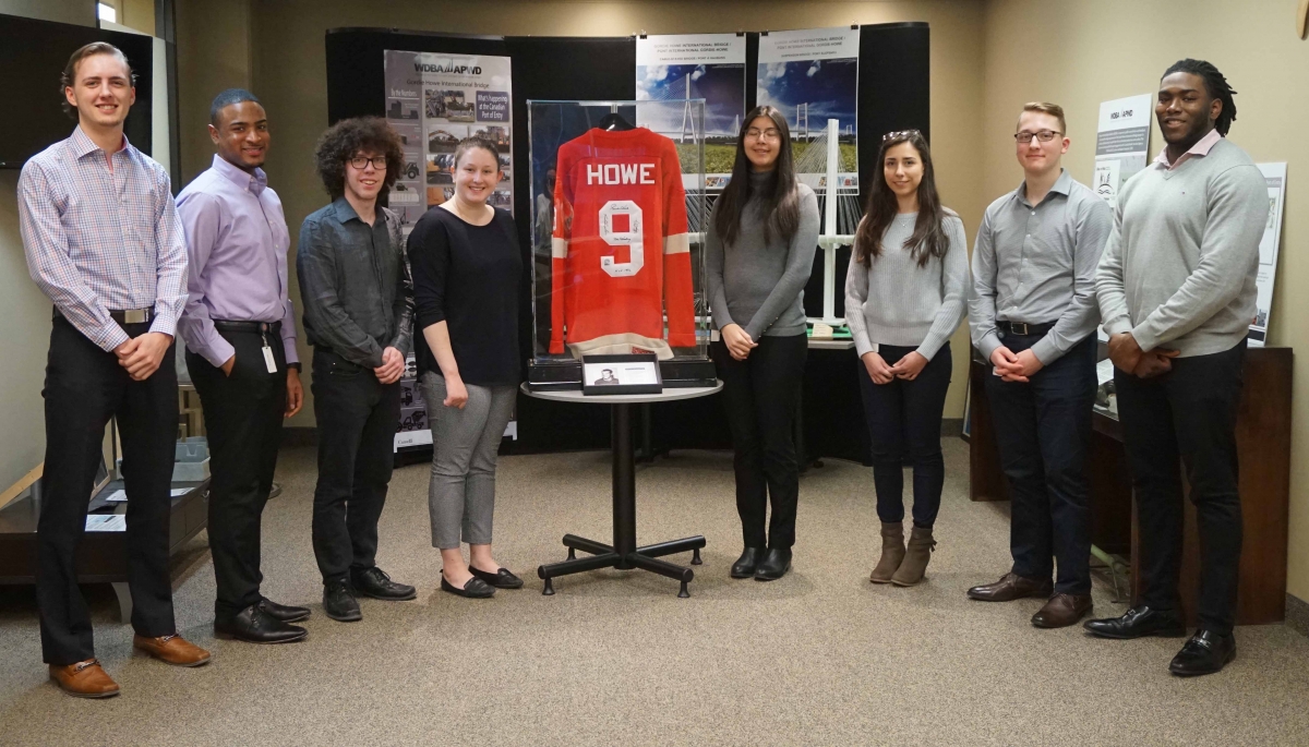 Photo of eight Co-Op Students at the WDBA Office in front of a Gordie Howe hockey jersey