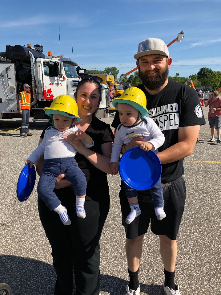 A couple holds their two young children standing outside in front of a large truck at the 2019 Meet-a-Machne event. 
